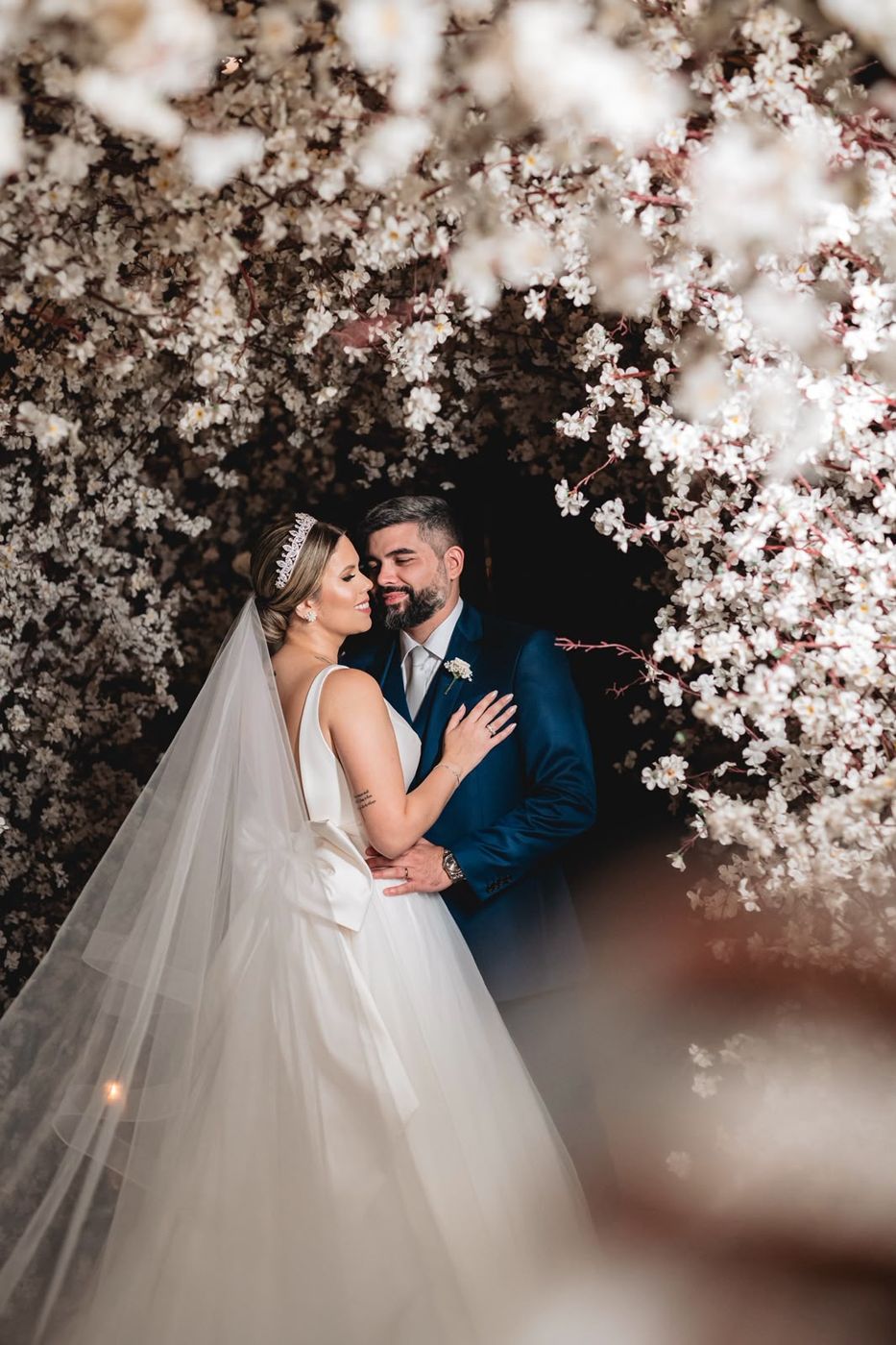 Noivos abraçados sob cascata de flores brancas em casamento assessorado por Dani Baida